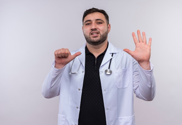 Young bearded male doctor wearing white coat with stethoscope showing and pointing up with fingers number six 