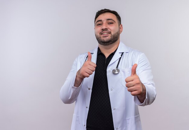 Молодой бородатый мужчина-врач в белом халате со стетоскопом показывает палец вверх с счастливой улыбкой на лице