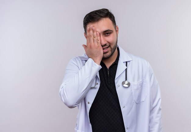Young bearded male doctor wearing white coat with stethoscope smiling covering one eye with palm 