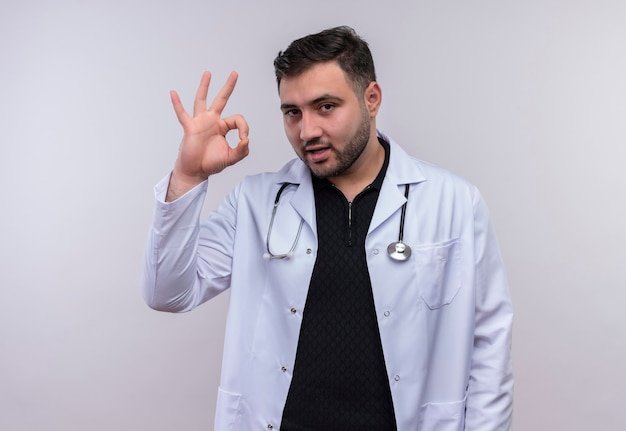 Young bearded male doctor wearing white coat with stethoscope smiling doing ok sign  over white  background
