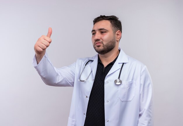 Молодой бородатый мужчина-врач в белом халате со стетоскопом улыбается, глядя в сторону, показывает палец вверх
