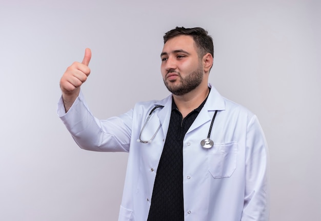 Young bearded male doctor wearing white coat with stethoscope smiling looking aside showing thumbs up 