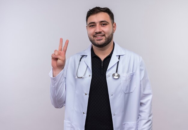 Young bearded male doctor wearing white coat with stethoscope smiling showing number two or victory sign 