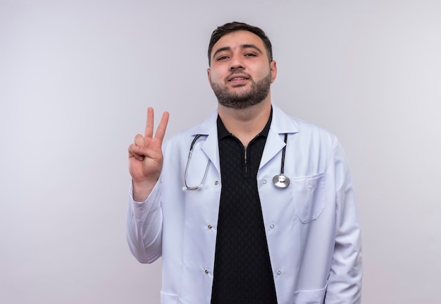 Young bearded male doctor wearing white coat with stethoscope smiling showing number two or victory sign 