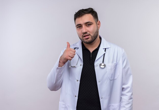 Young bearded male doctor wearing white coat with stethoscope smiling showing thumbs up 