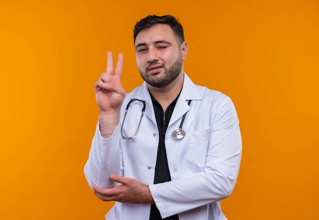 Молодой бородатый мужчина-врач в белом халате со стетоскопом подмигивает, улыбается и показывает знак победы