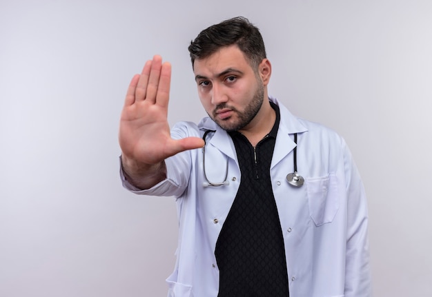 Молодой бородатый мужчина-врач в белом халате со стетоскопом с открытыми руками делает знак остановки с серьезным лицом