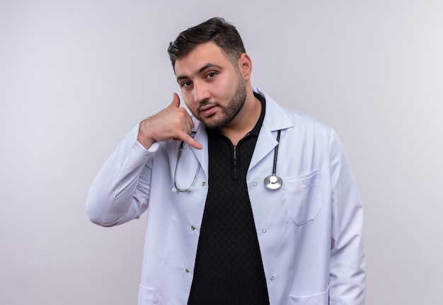 Young bearded male doctor wearing white coat with stethoscopelooking confident making call me gesture 