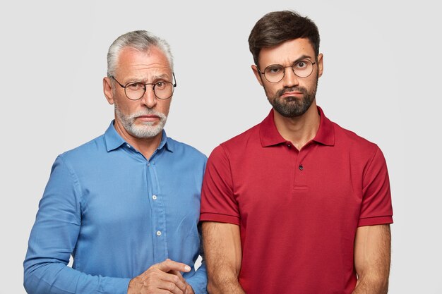 젊은 수염 난 남성은 당혹감에 눈썹을 올리고 빨간 티셔츠를 입고 성숙한 아버지 옆에 서서 흰 벽 위에 고립 된 가족 서클에서 주말을 보냅니다. 관계 개념
