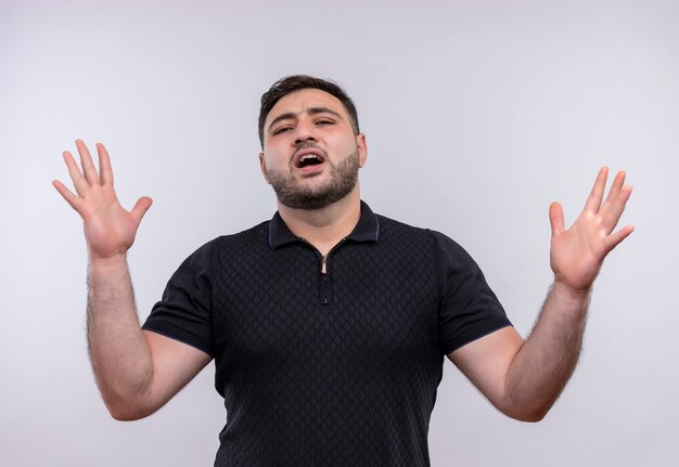Young bearded man in black shirt angry and frustrated with raised hands shouting 