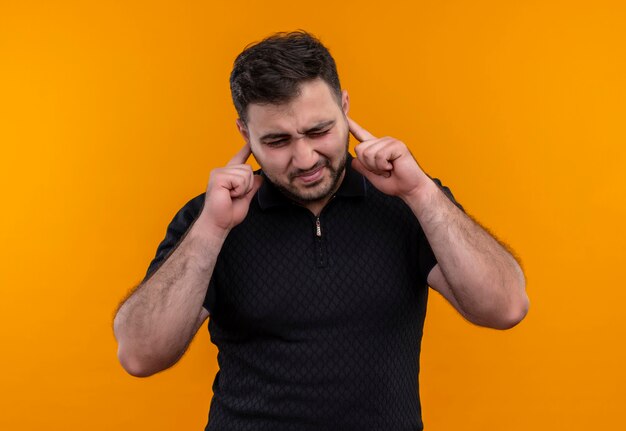 Young bearded man in black shirt  closing ears with fingers with annoyed expression 