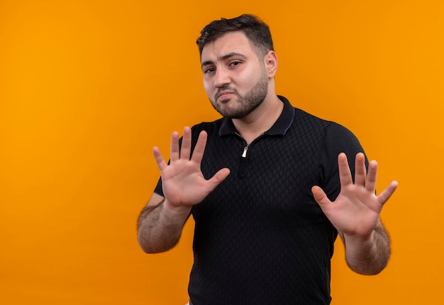 Young bearded man in black shirt   holding arms up making defense gesture looking confused 