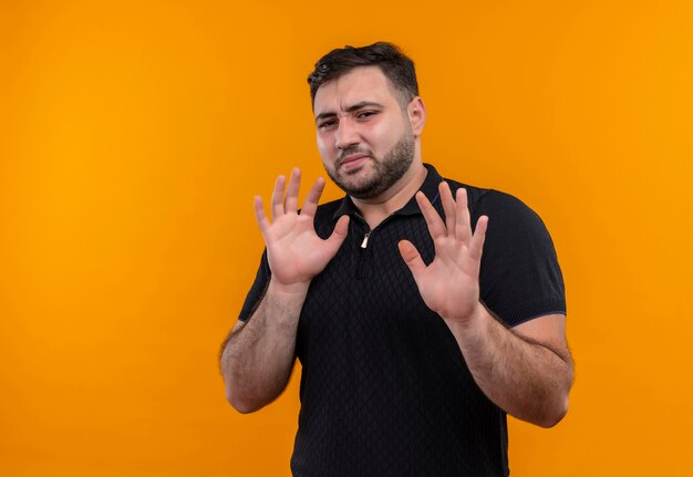 Young bearded man in black shirt   holding arms up making defense gesture with disguted expression 