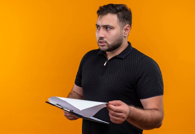 Young bearded man in black shirt  holding clipboard looking aside with confuse expression 