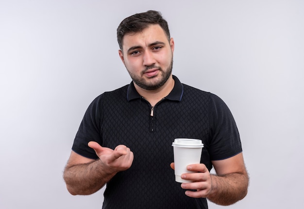 Young bearded man in black shirt holding coffee cup looking at camera displeased gesturing with hand 