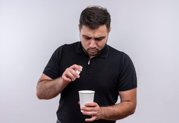 Young bearded man in black shirt holding coffee cup looking at it with serious face 