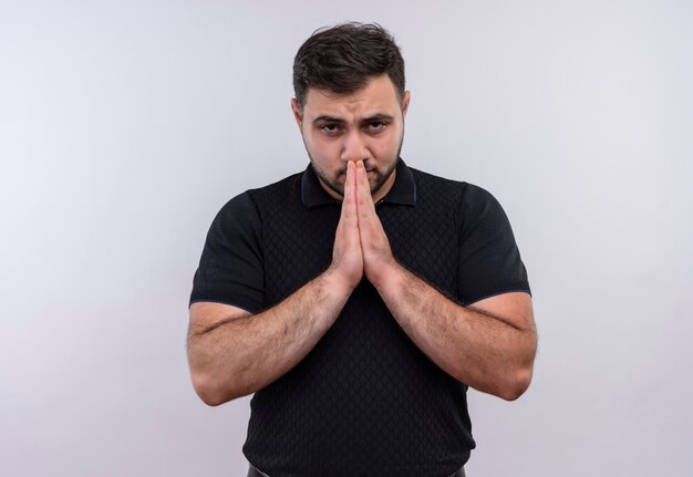Young bearded man in black shirt   holding hands like praying worried and emotional looking 