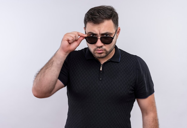 Young bearded man in black shirt holding  measure taking off his glasses looking at camera with frowning face 