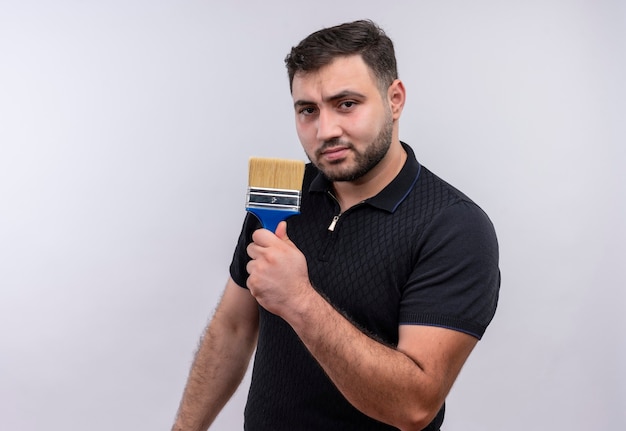 Young bearded man in black shirt holding  paint brush looking at camera with serious confident expression 