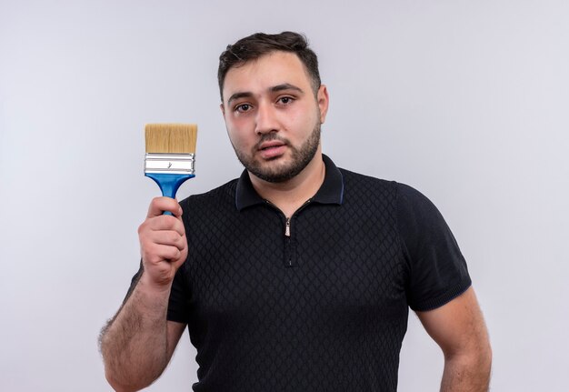 Free photo young bearded man in black shirt holding paint brush looking at camera with serious face