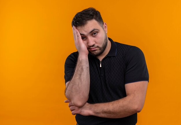 Young bearded man in black shirt  looking at camera leaning head on arm bored and tired 