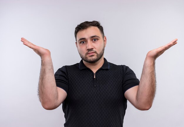 Young bearded man in black shirt looking confused and uncerating shrugging shoulders 