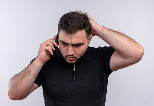 Молодой бородатый мужчина в черной рубашке выглядит смущенным и очень обеспокоенным во время разговора по мобильному телефону