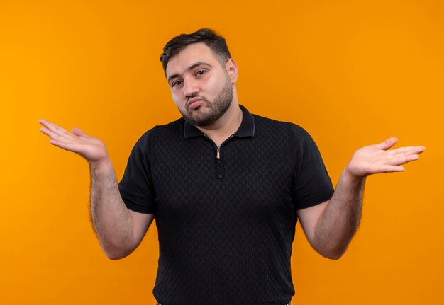 Young bearded man in black shirt looking uncertain and confused spreading arms to the sides having no answer 