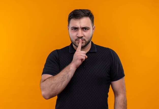 Young bearded man in black shirt  making silence gesture with finger on lips looking at camera with frowning face 