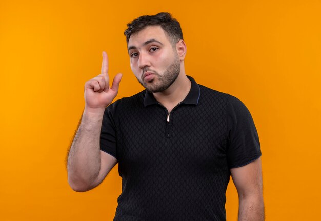 Young bearded man in black shirt  pointing with index finger up looking at camera with serious confident expression 