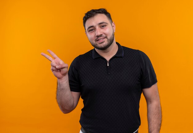 Young bearded man in black shirt  smiling cheerfully showing victory sign 