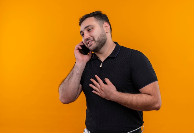 Молодой бородатый мужчина в черной рубашке улыбается счастливым и позитивным во время разговора по мобильному телефону