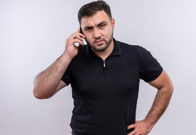 Молодой бородатый мужчина в черной рубашке разговаривает по мобильному телефону с сердитым лицом