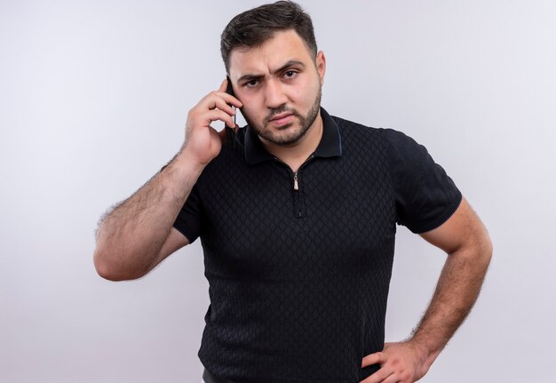 Young bearded man in black shirt talking on mobile phone with angry face 