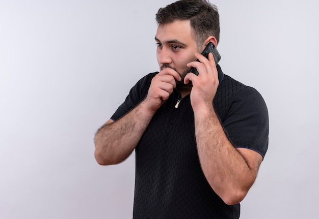 Молодой бородатый мужчина в черной рубашке рассказывает секрет во время разговора по мобильному телефону