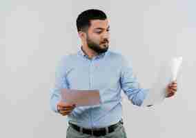 Free photo young bearded man in blue shirt holding blank pages looking at them with serious face standing over white wall