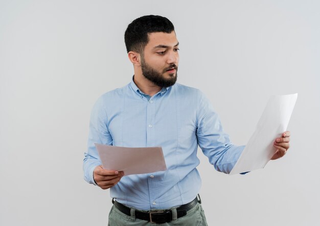 Young bearded man in blue shirt holding blank pages looking at them with serious face standing over white wall