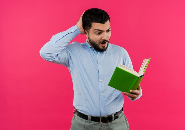 Молодой бородатый мужчина в синей рубашке держит книгу, глядя на нее удивленно и смущенно