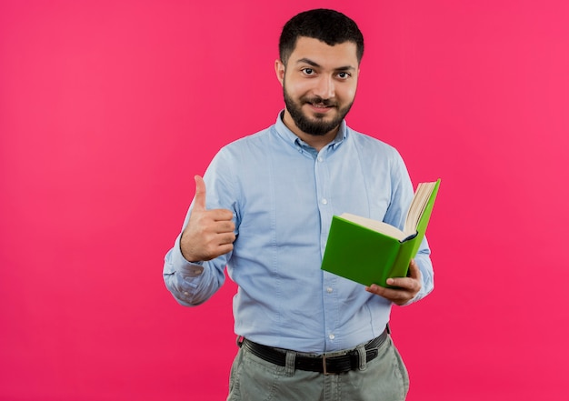 Молодой бородатый мужчина в синей рубашке держит книгу, улыбаясь, показывает палец вверх