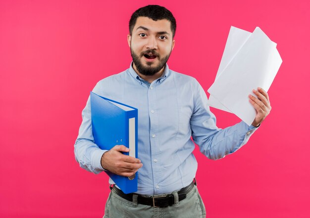 Young bearded man in blue shirt holding folder and blank pages happy and excited 