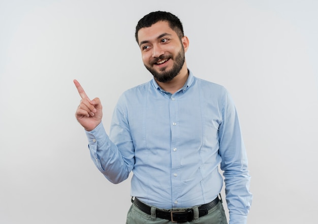 Young bearded man in blue shirt looking aside pointing with index finger to the side smiling 