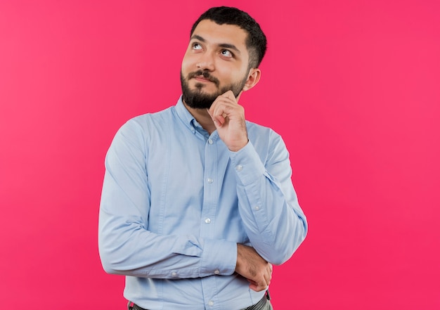 Giovane uomo barbuto in camicia blu che osserva in su con la mano sul mento pensando