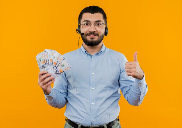 Young bearded man in blue shirt with headphones with microphone holding cash showing thumbs up smiling with happy face 