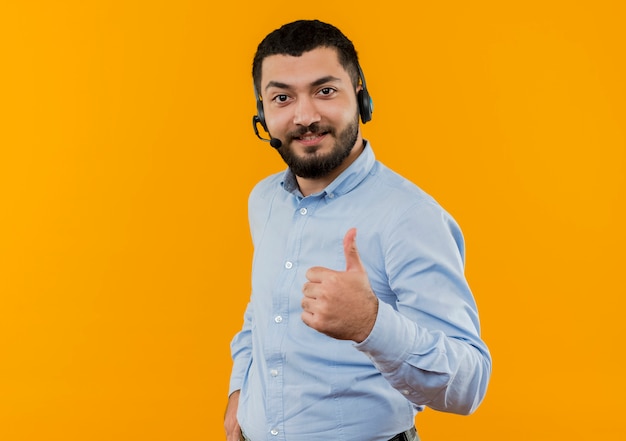 Foto gratuita giovane uomo barbuto in camicia blu con cuffie con microfono guardando davanti sorridente che mostra i pollici in su in piedi sopra la parete arancione