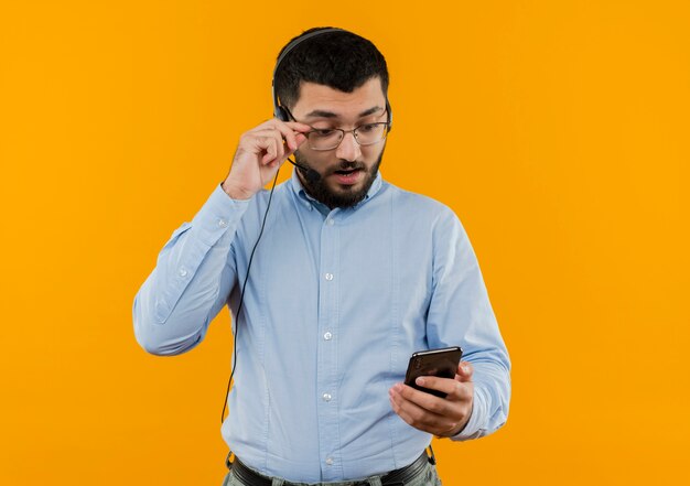 青いシャツを着た若いひげを生やした男が彼の眼鏡を外して彼のスマートフォンの画面を見ているマイクとヘッドフォン