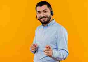 Free photo young bearded man in blue shirt with headphones with microphone pointign with index fingers at camera smiling cheerfully