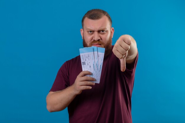 Young bearded man in brown t-shirt holding air tickets looking displeased showing thumbs down 