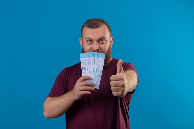 Young bearded man in brown t-shirt holding air tickets showing thumbs up 