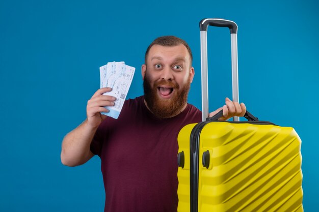 Молодой бородатый мужчина в коричневой футболке с дорожным чемоданом и авиабилетами выглядит возбужденным и счастливым