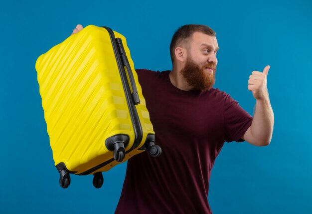 Молодой бородатый мужчина в коричневой футболке держит дорожный чемодан, улыбаясь, указывая пальцем назад
